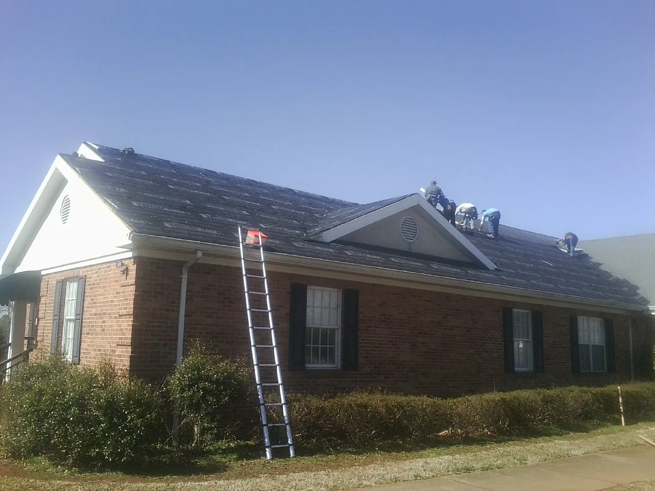 Crew tearing shingles off a brick ranch roof with a ladder set — Emergency Roof Repair work by our crew in Sandy Spring