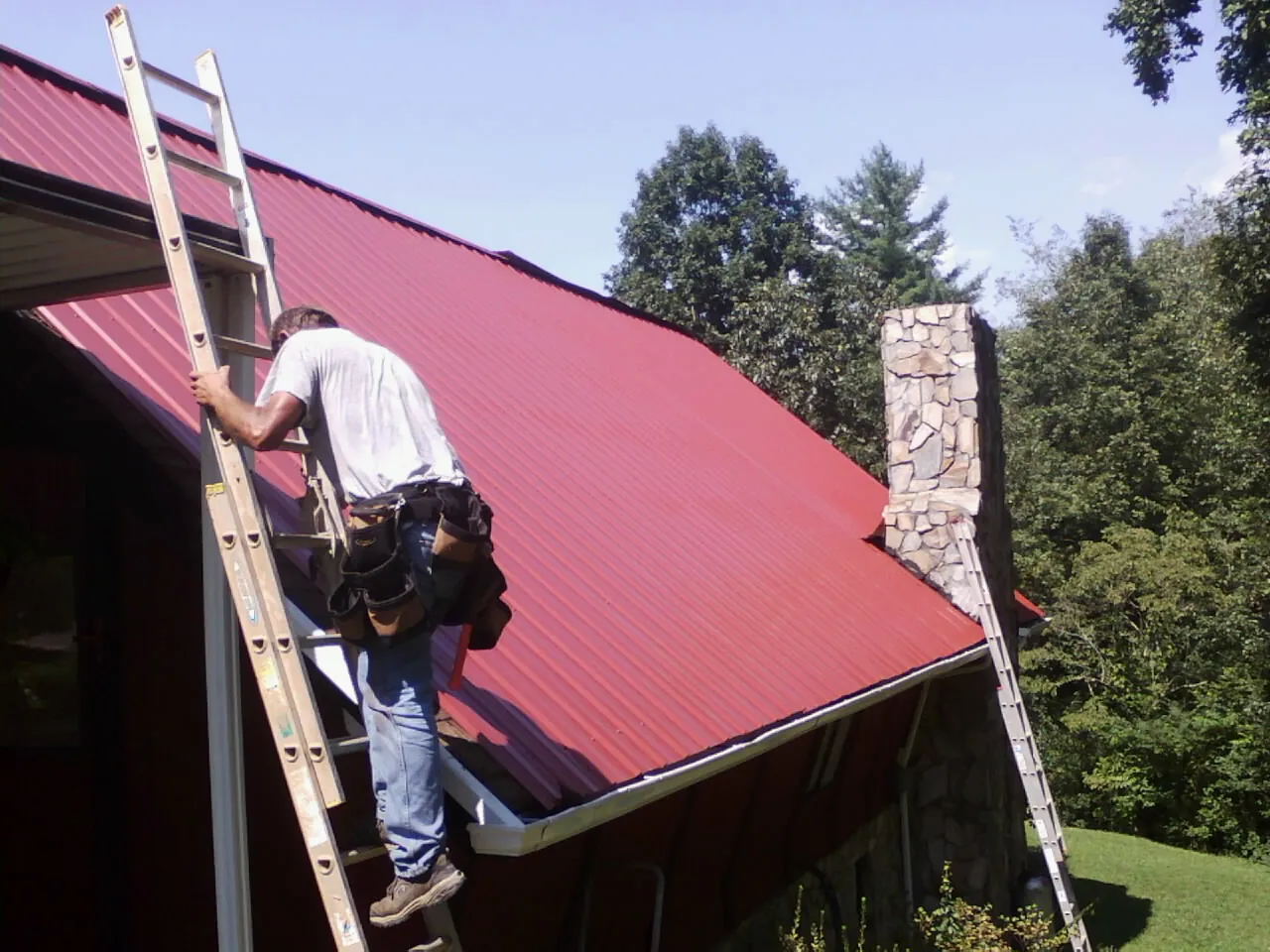 Roofer on a ladder beside a red metal roof and stone chimney — Soffit & Fascia Repair by Roofing Brothers in Sandy Spring